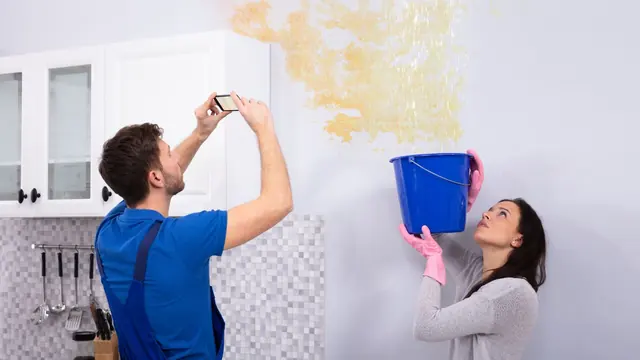 Renter holding a bucket under a leak while a man photographs the stained kitchen wall